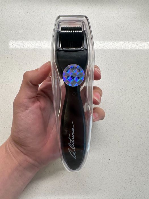 A hand holding a derma roller in a clear plastic case. The black handle has "Altima" written on it and a holographic sticker shows "0.5mm."